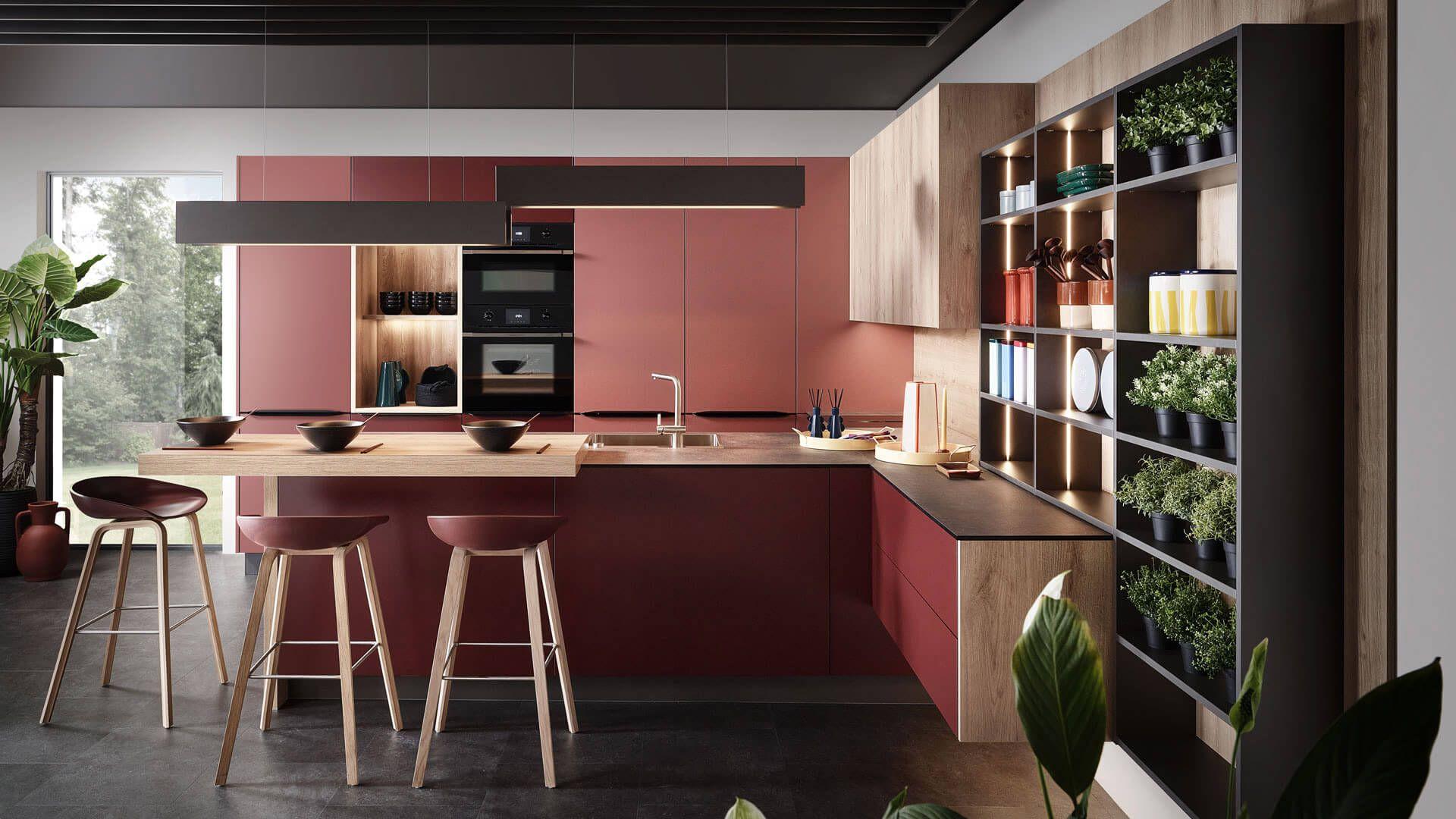 Moderne Sachsenküche (Modelle Selma und Lana) in Terrakotta-Rot und warmer Holz-Optik, präsentiert vom Küchenstudio Borcherts in Berlin. Einbauküche in L-Form mit integrierter Frühstückstheke, Barhockern und einem offenen, beleuchteten Design-Regal.
